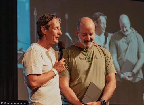 Two men on stage, one with microphone resting hand on the other's shoulder, large projected image behind them