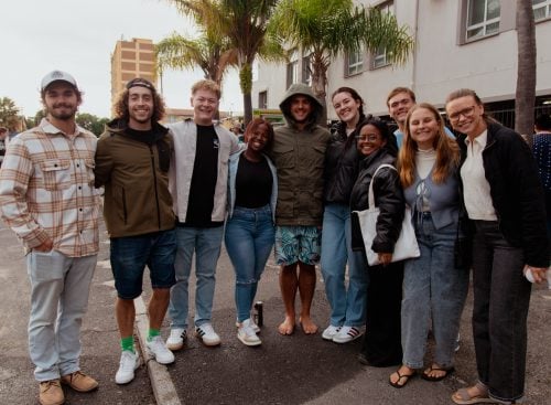 Group of ten people standing close together on a street, smiling with palm trees and a building behind