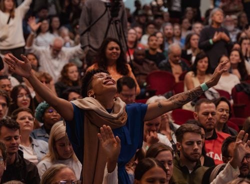 Large seated congregation with a woman in the centre standing and raising her arms in worship at a conference