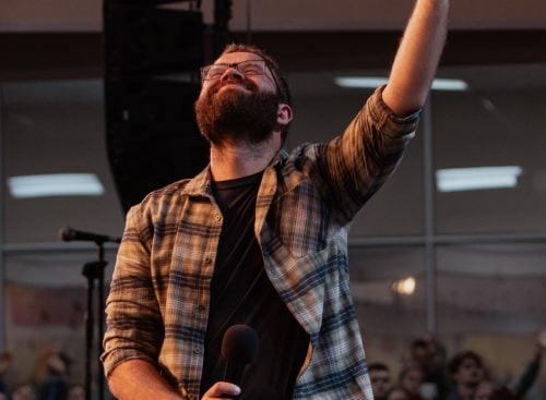 Bearded man kneeling on stage holding a microphone with one hand raised, audience visible in the background