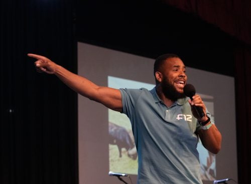 Male speaker in a blue Four12 polo shirt holding a microphone and pointing on stage with a projection screen behind him
