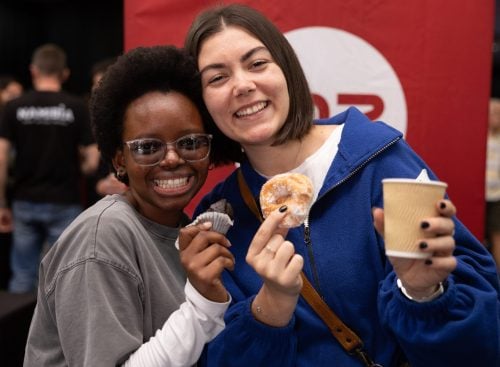 Two young people smiling at camera, one holding a doughnut and takeaway coffee, the other holding a cupcake at an indoor