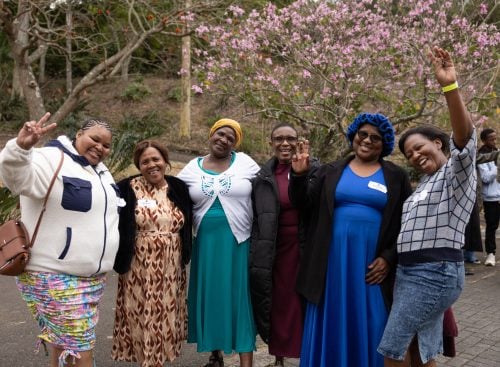 Six women standing together outdoors in front of a flowering tree, smiling and waving at the camera