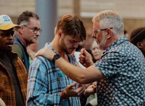 Older man with hand on a young man's shoulder while praying in a group setting
