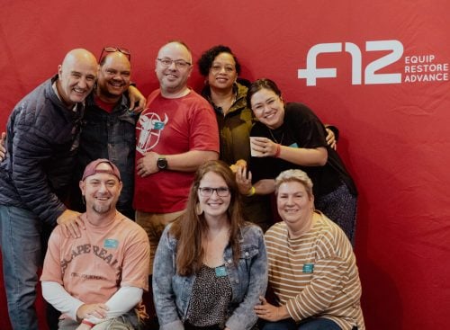 Eight people smiling and posing in front of a red F12 backdrop reading 'Equip Restore Advance'