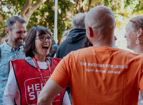 Group of people outdoors talking; a woman in a red volunteer vest laughing and a man in an orange T‑shirt seen from behind