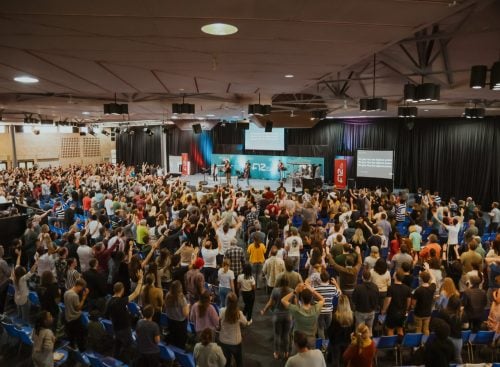 Large congregation standing in a hall facing a stage with a speaker and musicians