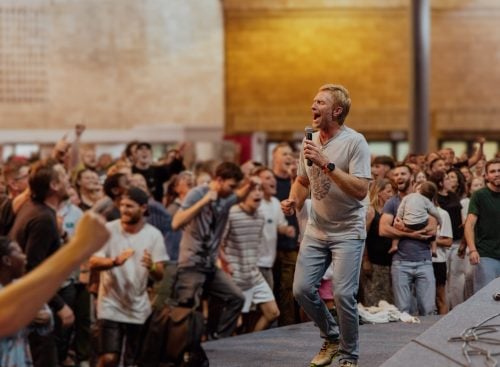 Male speaker holding a microphone on stage leading an enthusiastic indoor crowd