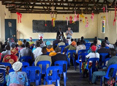 Congregation seated on blue plastic chairs facing two speakers at the front of a decorated church hall