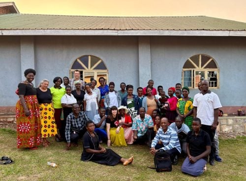 Large mixed-age community group posed outside a single-storey building with arched windows