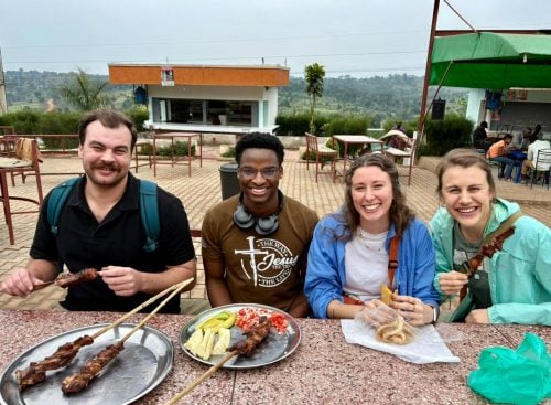 Four people seated at an outdoor table eating skewered meat and snacks with a countryside view